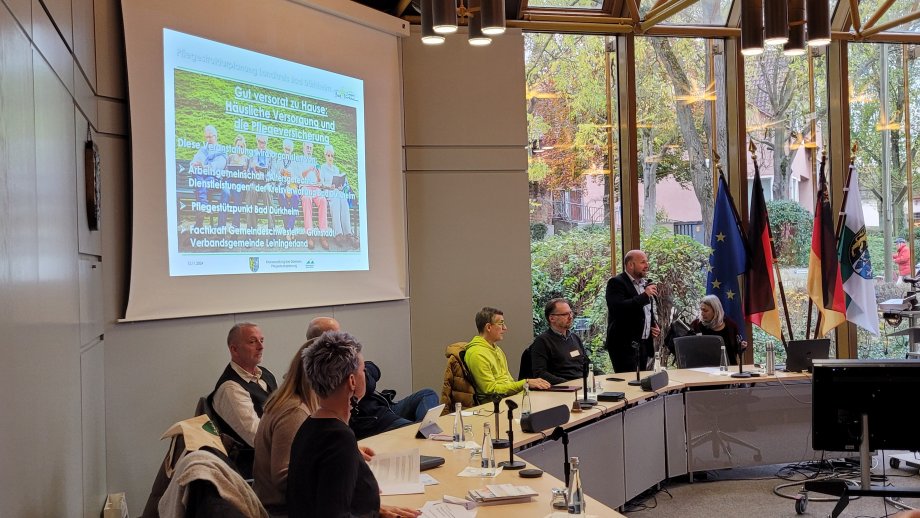 Ein Mann spricht in ein Mikrofon. Daneben sitzen mehrere Menschen. Im Hintergrund eine Präsentation auf einer Leinwand.