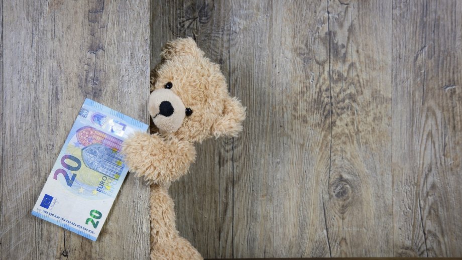 Ein Teddy hält einen 20-Euro-Schein mit einer Pfote vor einer Holzwand fest.