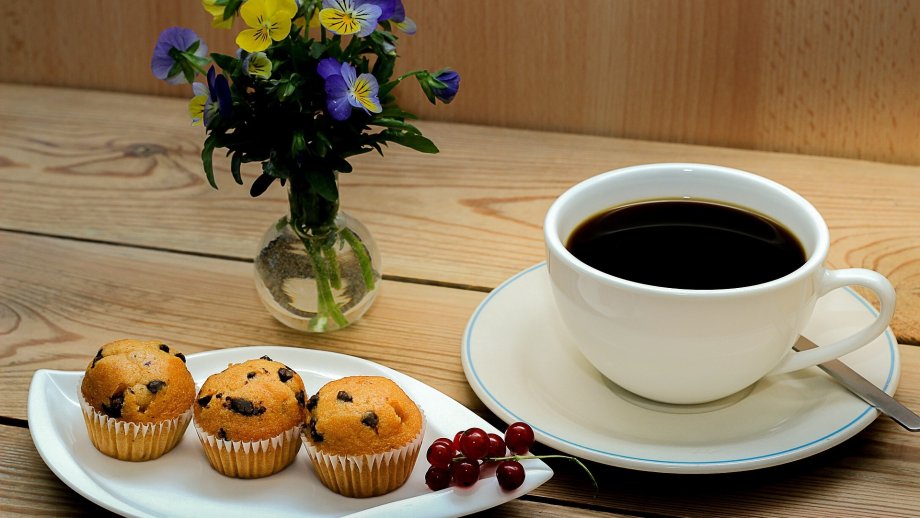 Eine Tasse Kaffee steht neben einem Teller mit drei Muffins, dahinter ein kleiner Blumenstrauß in einer Vase.