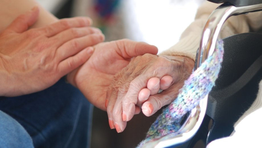 Zwei junge Hände halten eine Hand einer älteren Frau mit zartrosa lackierten Fingernägeln. 
